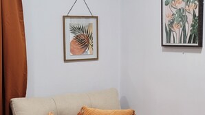 En beige soffa med två orange kuddar, en inramad växt på väggen och en brun gardin.