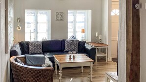 Ein Wohnzimmer mit einer Couch, einem Couchtisch und einem Korbstuhl. Zwei Fenster mit Jalousien, eine Lampe und ein kleiner Tisch mit Kerzen.