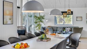 Un comedor moderno con una mesa larga, sillas negras y un frutero. Dos lámparas colgantes iluminan la mesa y se ve un ventanal grande al fondo.