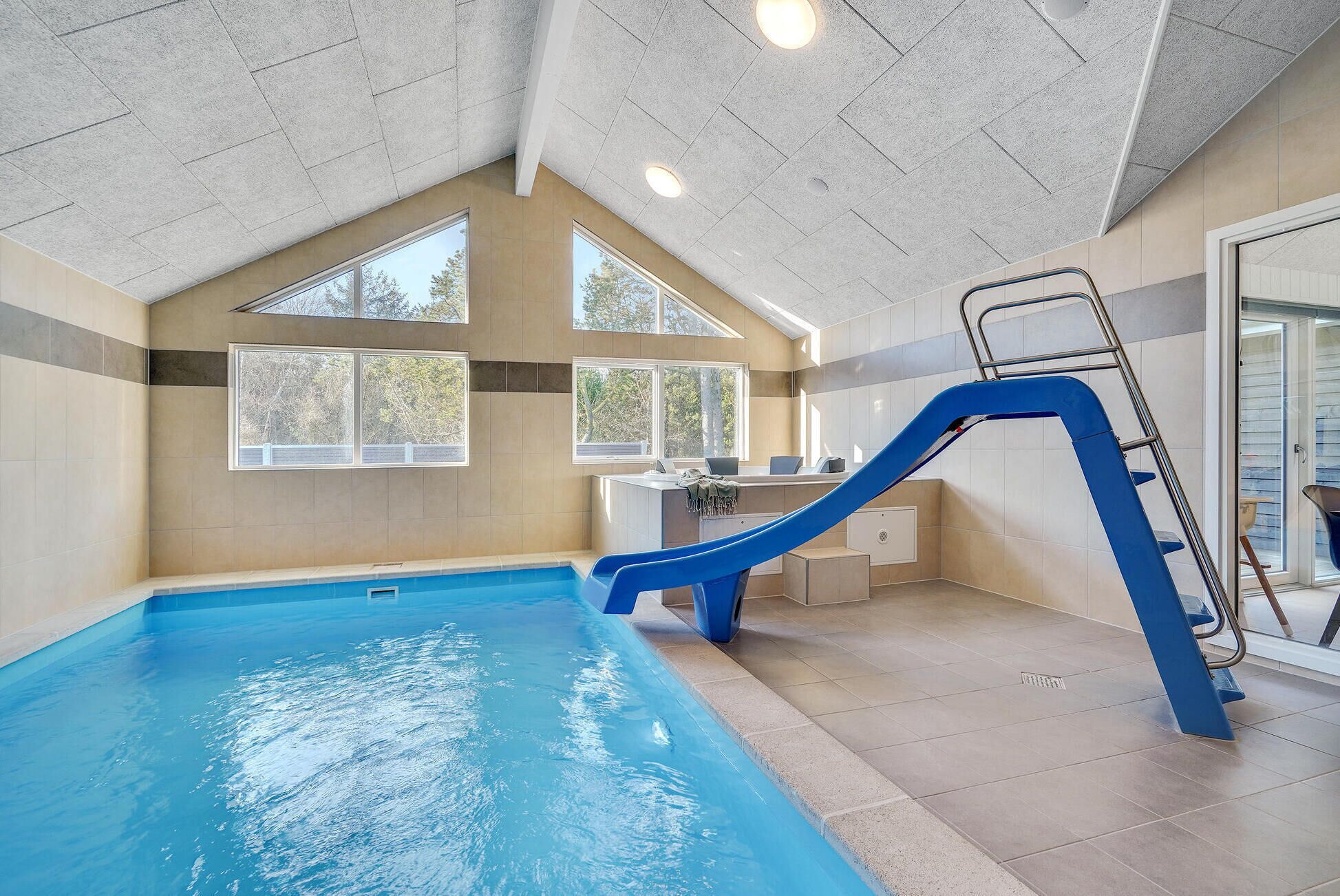An indoor swimming pool with a blue slide and a ladder, large windows, and a modern interior design.
