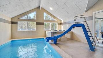 An indoor swimming pool with a blue slide and a ladder, large windows, and a modern interior design.