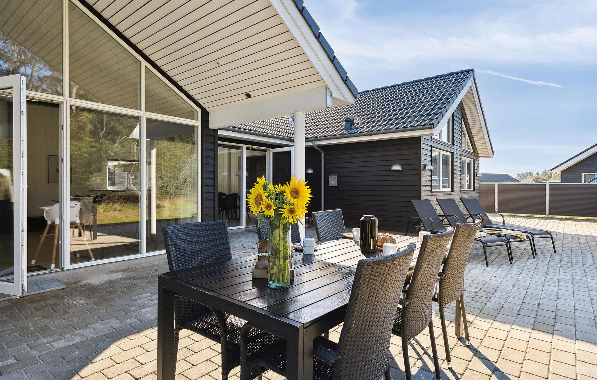 A modern house with a patio area featuring a table and chairs, a large window, and a view of the surrounding landscape.