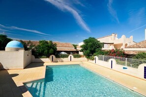 A swimming pool with clear blue water, surrounded by a white wall with blue accents and a tiled roof structure in the background.