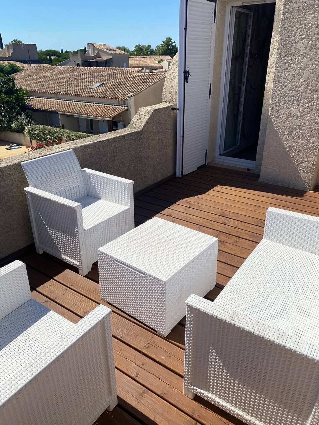 A rooftop terrace with white wicker furniture, a wooden deck, and a view of surrounding buildings.