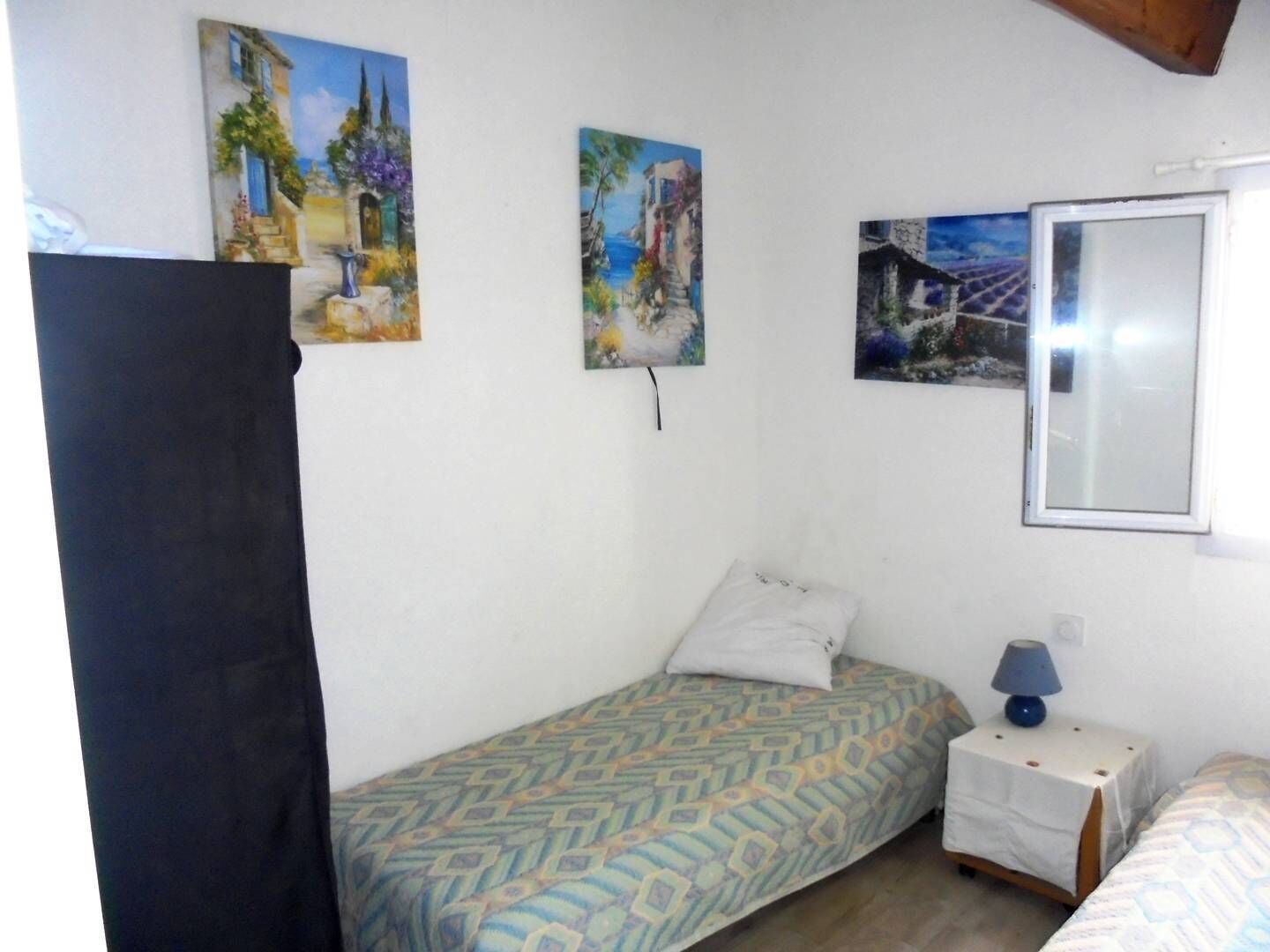 A bedroom with two beds, a small bedside table with a lamp, and three paintings on the wall.