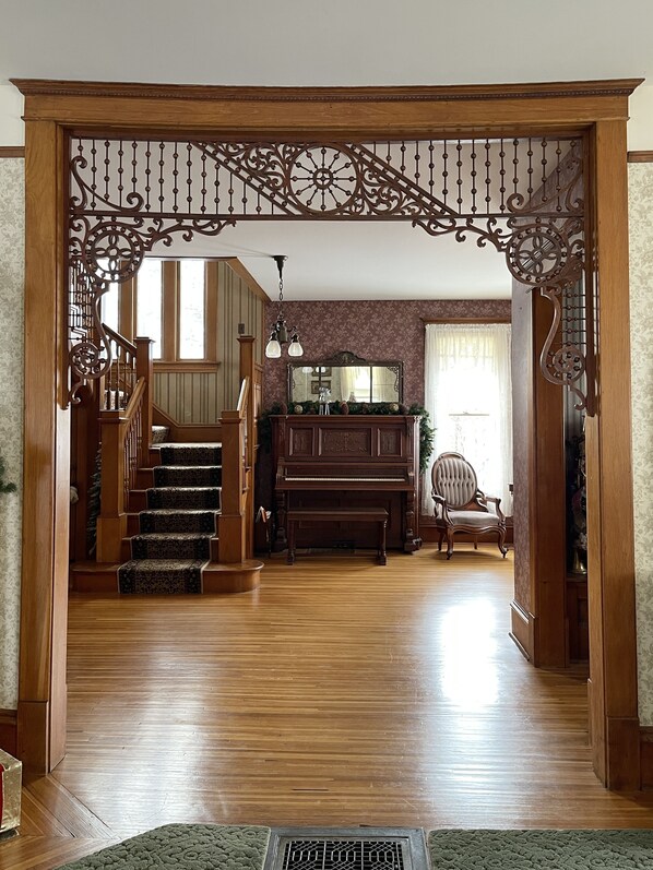 Une entrée imposante avec un escalier en bois menant à un salon agrémenté d’un piano, d’un grand miroir et d’un fauteuil décoratif.