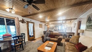 A cozy living room with a wooden ceiling, a dining area with a glass table, and a kitchen in the background. - Croll Cabins-Armstrong Library- #5 at Vallecito Lake, private lawn, great views! (Bayfield)