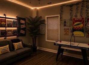 A cozy room with a pool table, a couch with decorative pillows, a potted plant, and various posters on the walls.