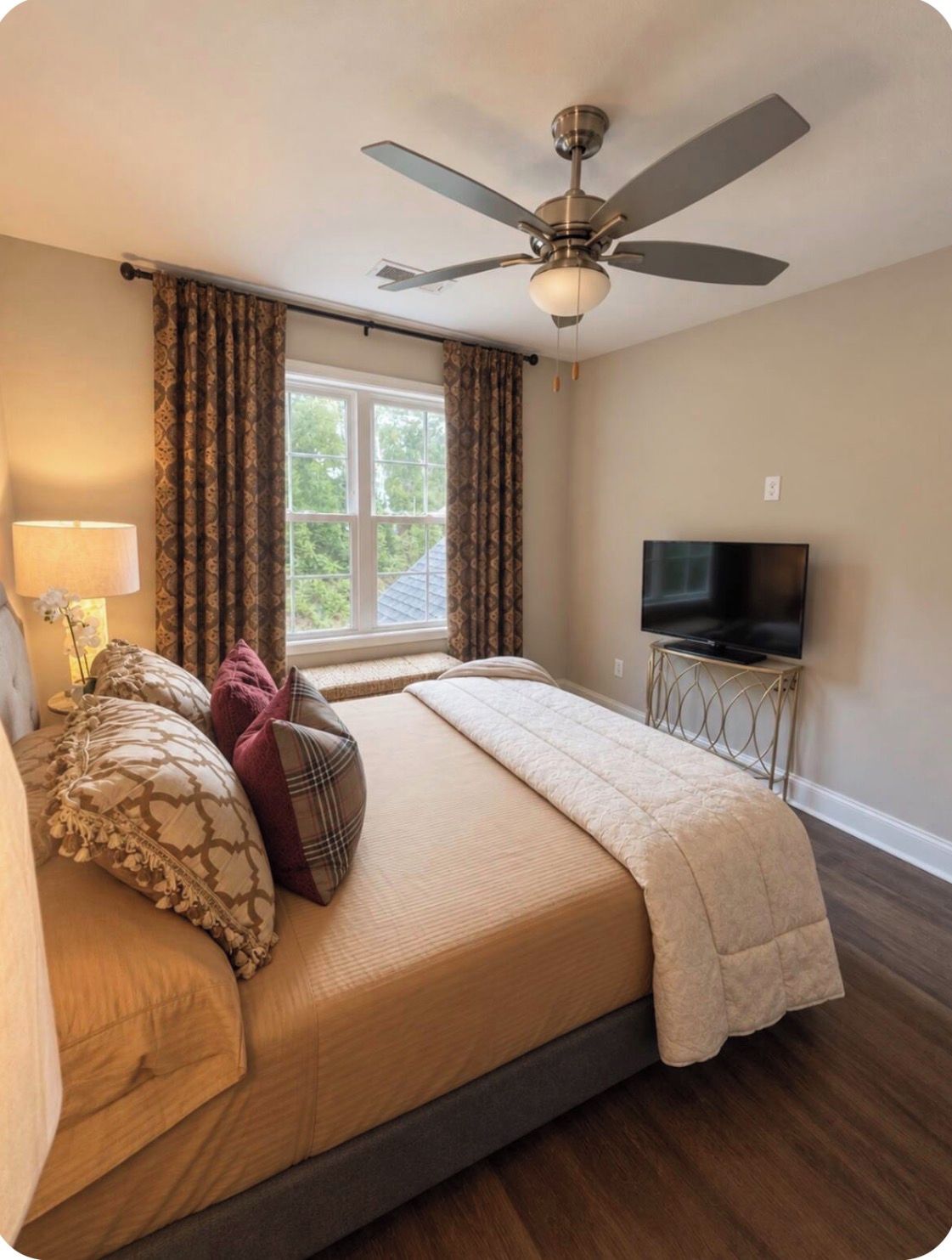 A bedroom with a bed, curtains, a ceiling fan, a television on a stand, and a window.