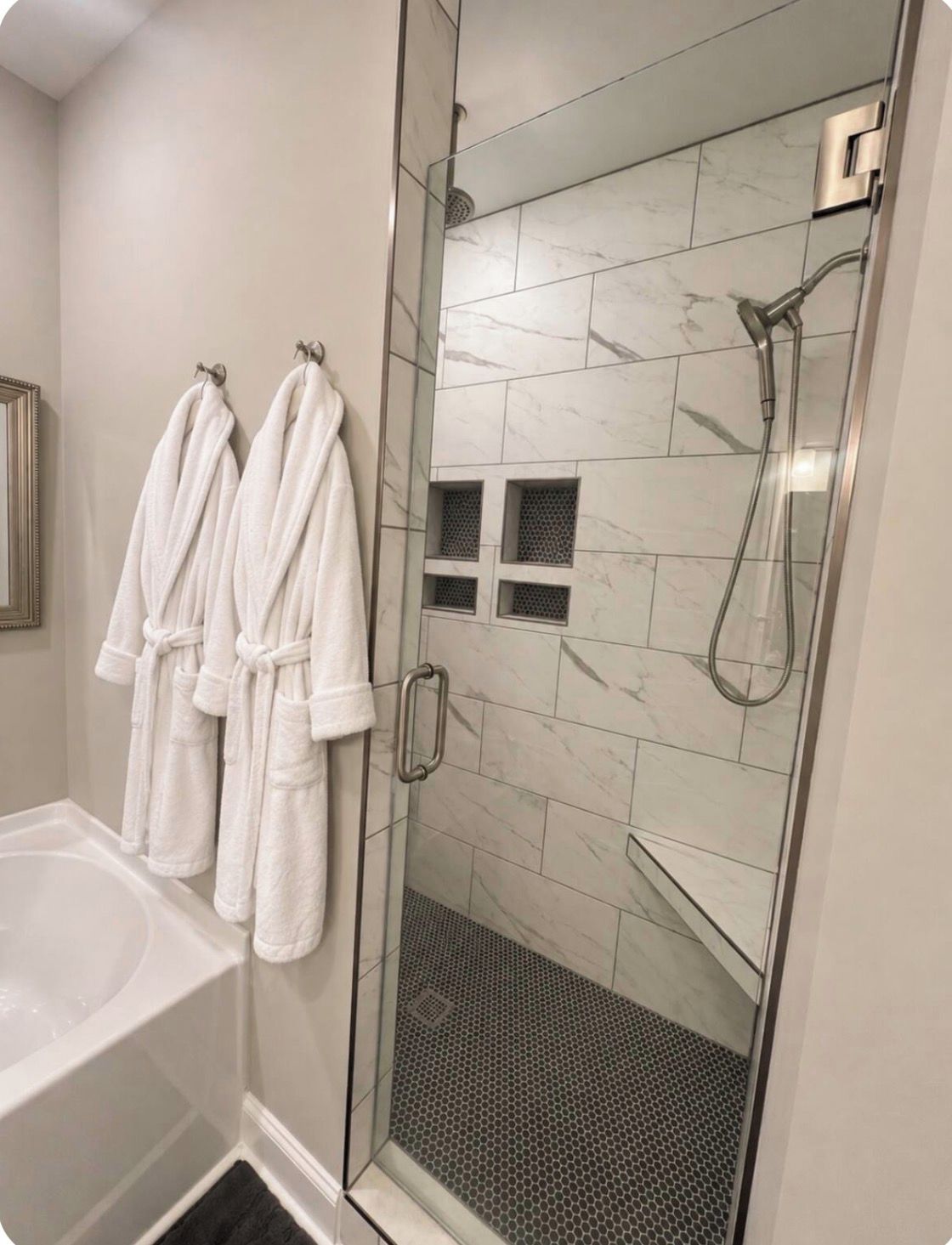 Un baño con ducha envuelta en vidrio, una bañera y dos albornoces blancos colgados en ganchos.