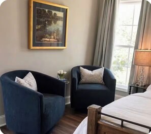 A hotel room with two armchairs, a bed, a nightstand with a lamp, and a framed picture on the wall.
