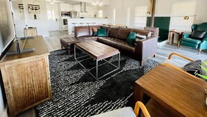 Een ruime woonkamer met een bruine leren bank, een houten salontafel en een zwart-wit tapijt. Er is een eethoek met een houten tafel en stoelen, en een keuken op de achtergrond.
