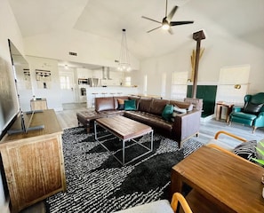 A spacious living room with a brown leather sofa, a wooden coffee table, and a black and white patterned rug. There is a dining area with a wooden table and chairs, and a kitchen in the background.