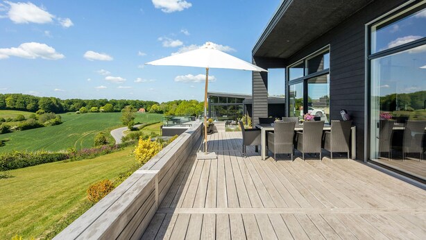A wooden deck with outdoor furniture and a large umbrella, overlooking a green landscape and a modern building with large windows. - 3 bedroom awesome home in Aabenraa (Aabenraa)
