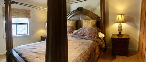 A four-poster bed with a patterned bedspread, a wooden nightstand with a lamp, and a window with a view of trees.