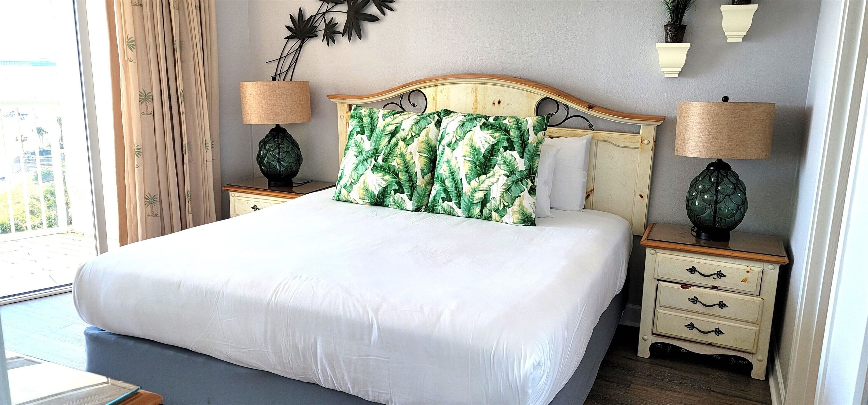 Uma cama bem arrumada com um edredom branco, um travesseiro estampado com folhagens tropicais e duas luminárias de cabeceira sobre mesas de cabeceira de madeira.