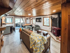 A cozy cabin interior with a wooden ceiling, a dining area, a sofa with a blanket, and a fireplace.