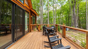 A wooden deck with a table and chairs, surrounded by a metal railing, overlooking a forested area. - Stillness Springs at Eagles Nest - Pet Friendly, Two master suites, firepit, hot tub, creek, 1 acre! (Banner Elk)