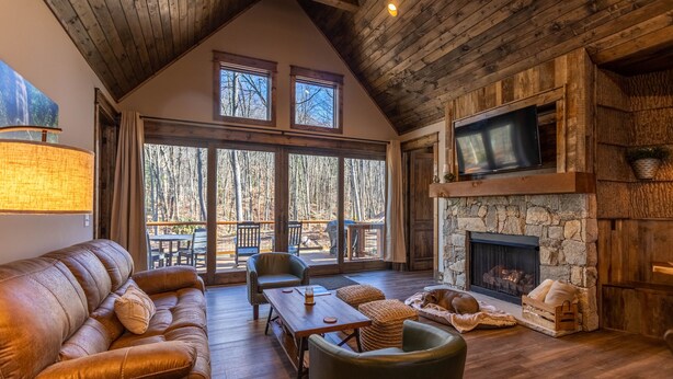 A cozy cabin interior with a stone fireplace, leather sofa, and a large window overlooking a forest. - Stillness Springs at Eagles Nest - Pet Friendly, Two master suites, firepit, hot tub, creek, 1 acre! (Banner Elk)