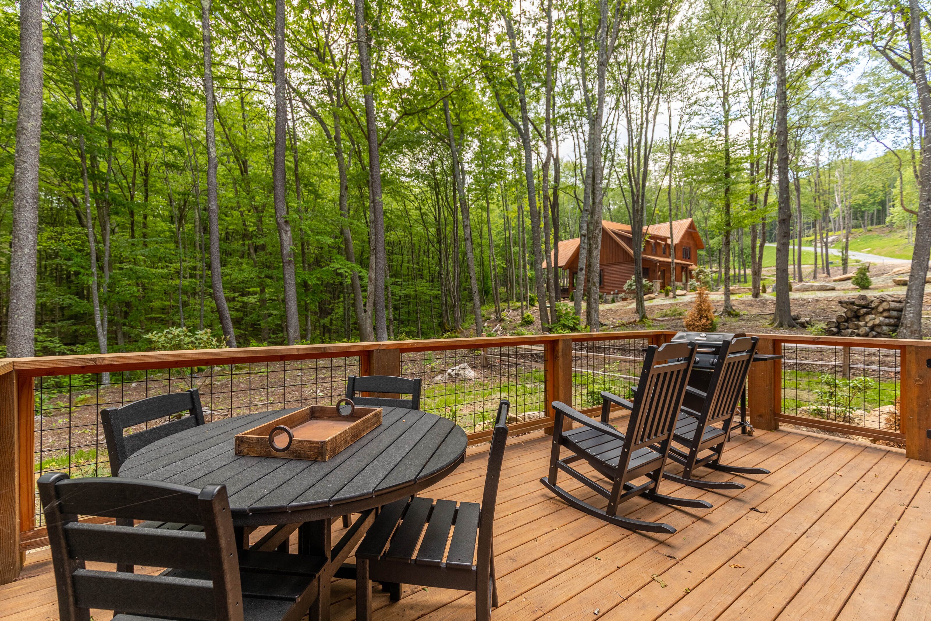 Eine Holzterrasse mit rundem Tisch und Stühlen, umgeben von einem Wald.