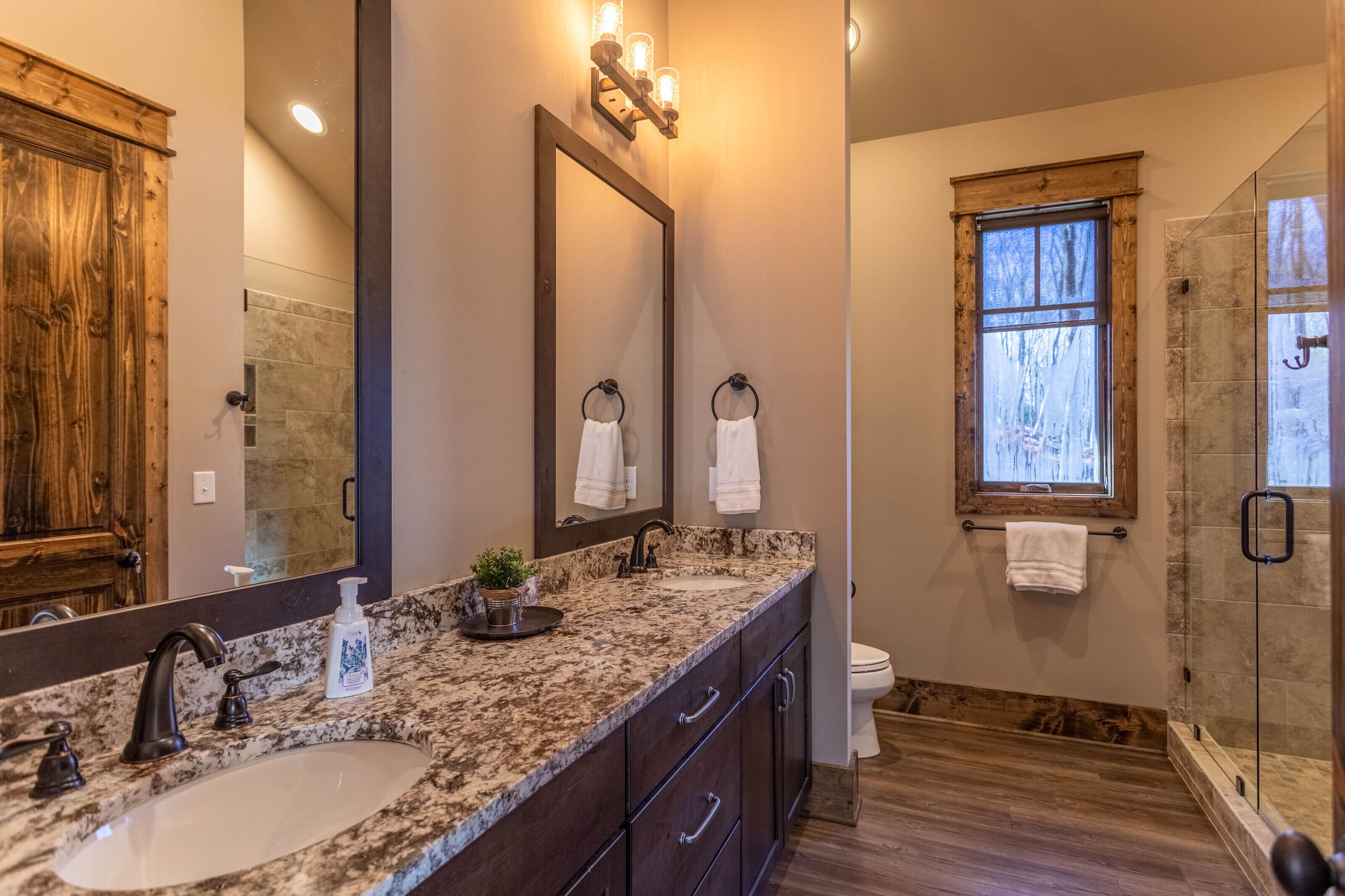 A bathroom with a granite countertop, a large mirror, a shower with a glass door, and a window providing natural light.