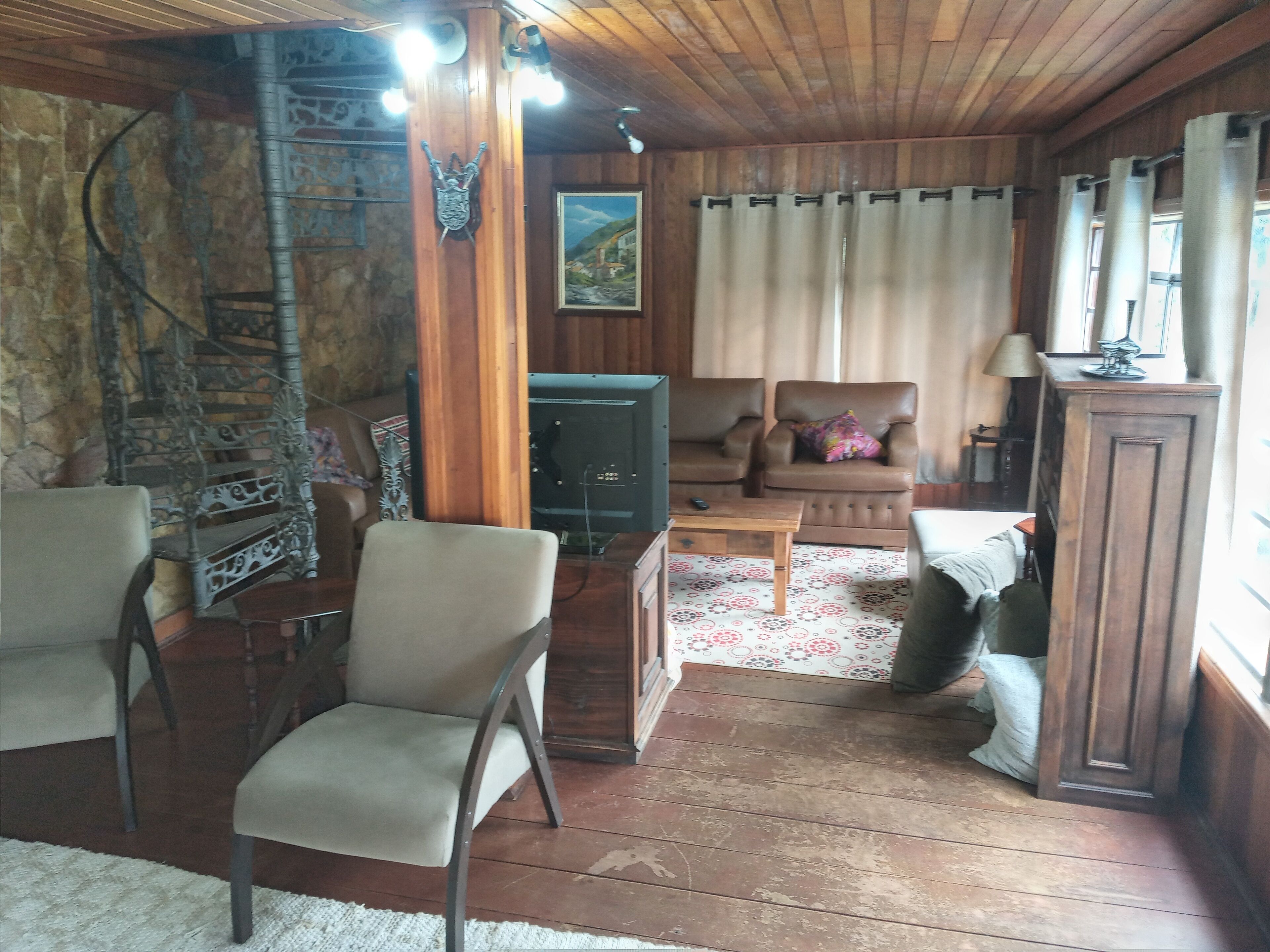 Un salon chaleureux avec un plafond en bois, un mur en pierre, un escalier, une télévision, un fauteuil en cuir marron, un fauteuil beige, un tapis à motifs et une fenêtre avec des rideaux.