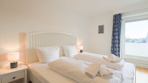 A hotel room with a bed, two bedside tables, a window with curtains, and a view of the outside. - Friesennerz - Hedighuis - Friesennerz (Sylt)
