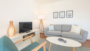 A modern living room with a grey sofa, a flat-screen TV, a small white table, and a floor lamp. - Friesennerz - Hedighuis - Friesennerz (Sylt)