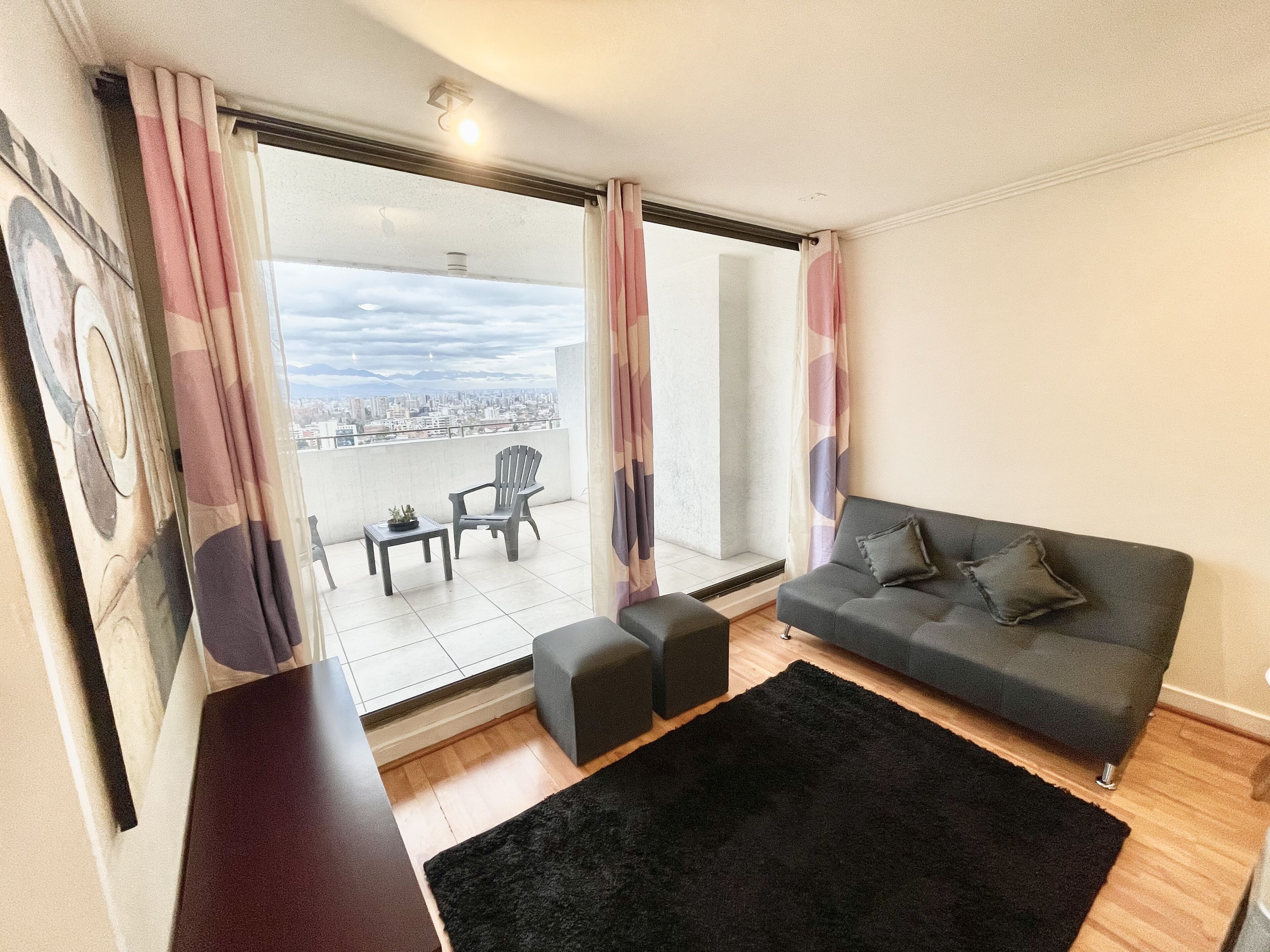 A living room with a grey sofa, a black rug, a small table with a plant, and a chair. There is a large window with curtains, and a view of a cityscape and mountains in the background.