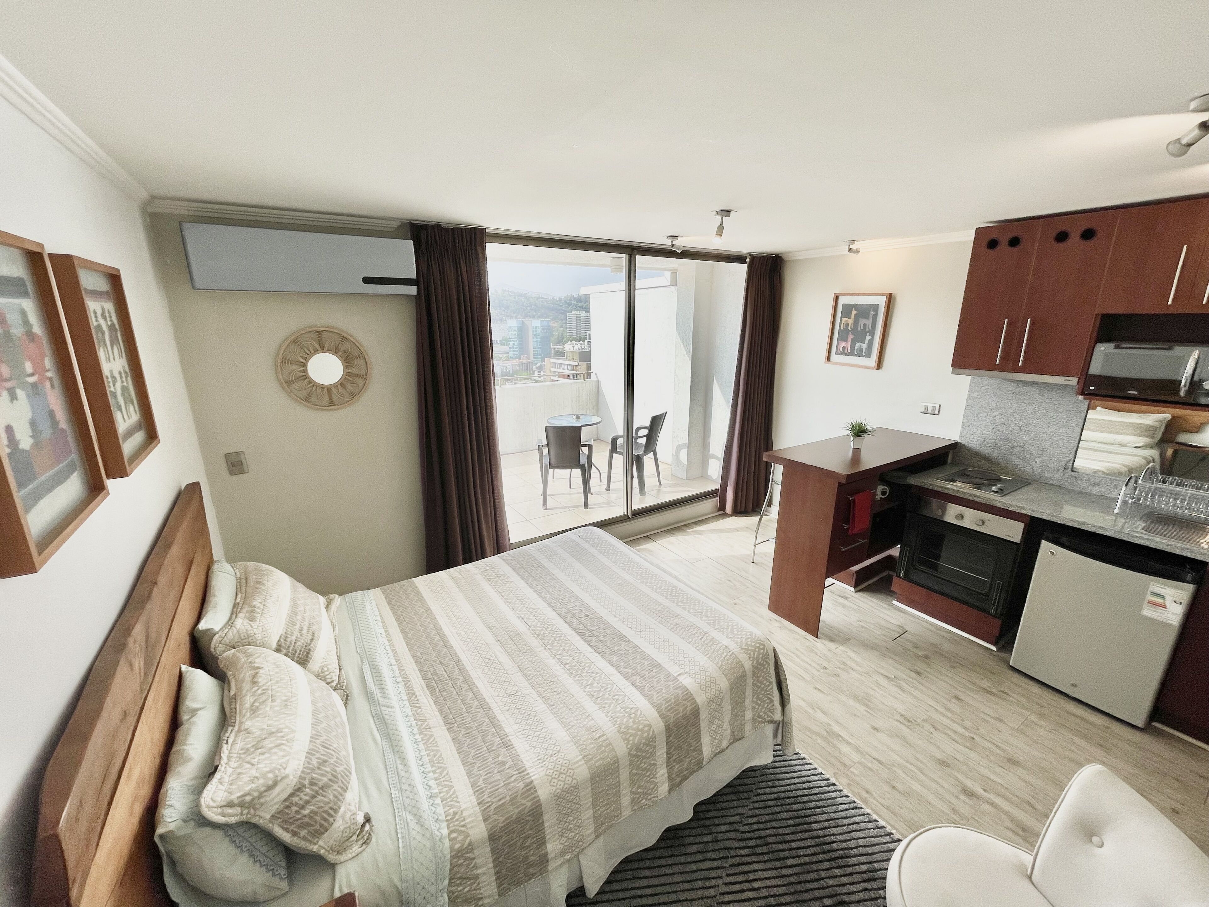 A hotel room with a bed, a small kitchenette, a dining table, and a balcony with a view.