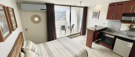 A hotel room with a bed, a small kitchenette, a dining table, and a balcony with a view.