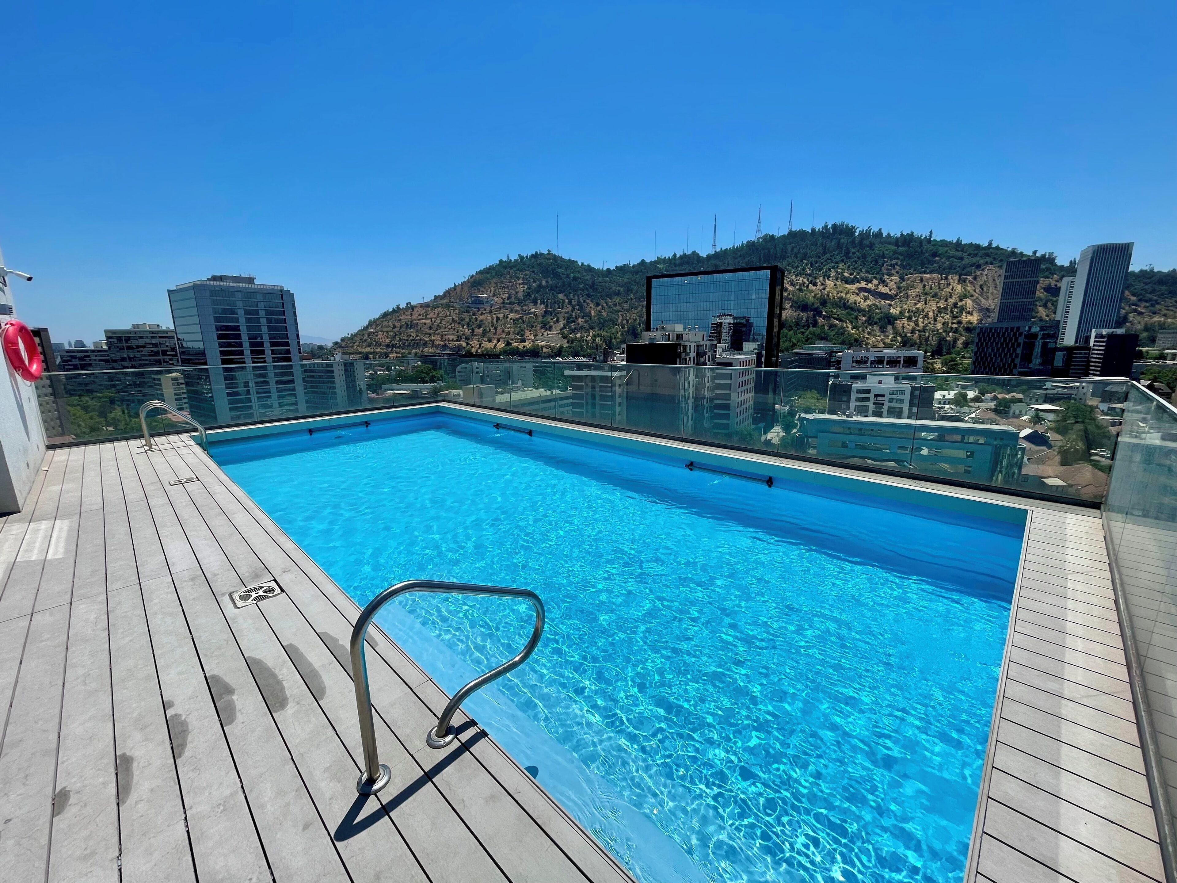 A rooftop pool with clear blue water, surrounded by a wooden deck and glass railing.
