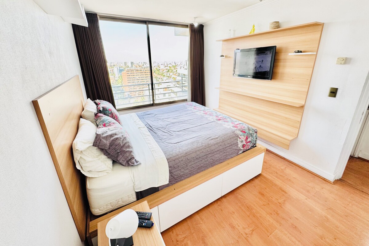 Un dormitorio moderno con una cama, un televisor montado en la pared y un ventanal con vista a la ciudad.