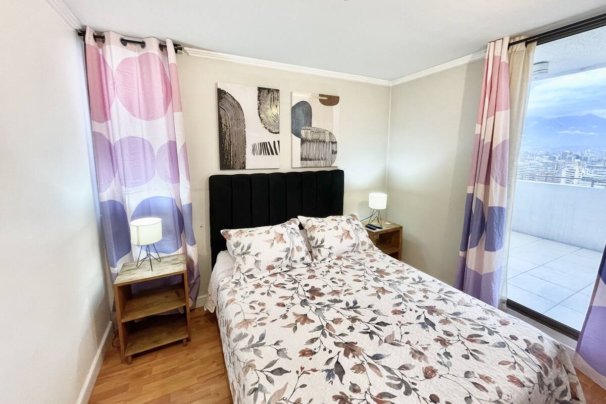A bedroom with a bed, a nightstand, a wall-mounted lamp, and a view of the city through the window.