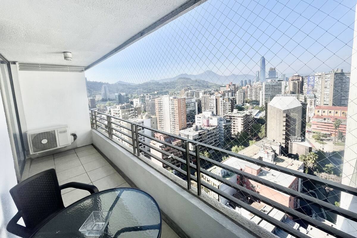 A balcony with a glass table and chairs, overlooking a cityscape with various buildings and a mountain in the distance.