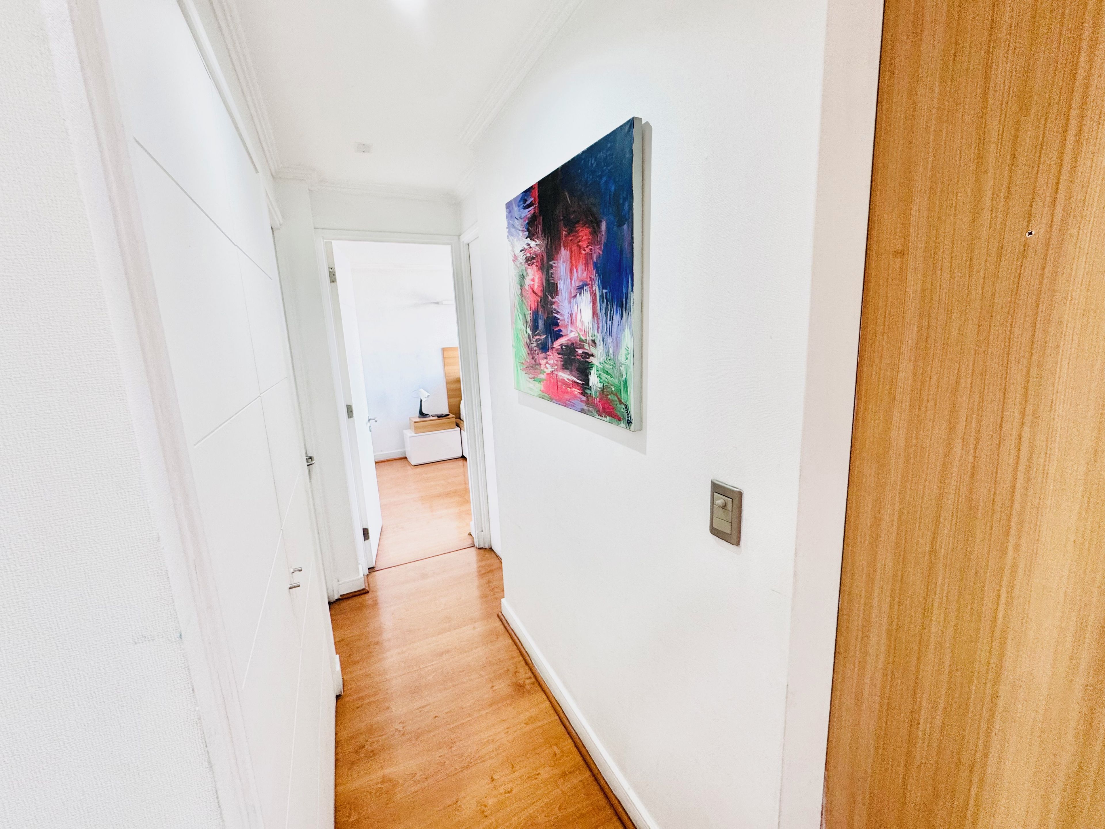 A hallway with white walls and wooden flooring, leading to a room with a bed and a nightstand.