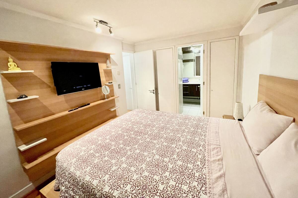 A bedroom with a bed, a TV mounted on a wooden shelf, a mirror, and a wardrobe.