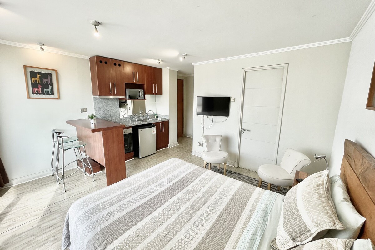 A compact hotel room with a bed, a small kitchenette, a TV mounted on the wall, and a bathroom.
