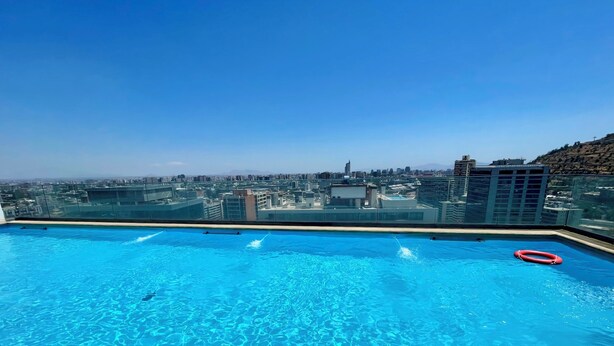 Una piscina en la azotea con agua cristalina azul, rodeada por una barandilla de vidrio, con vistas a la ciudad.