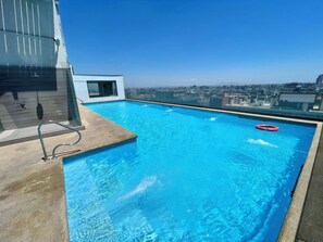 A rooftop swimming pool with clear blue water, a glass balustrade, and a city view in the background. - Rho.cl (Santiago)
