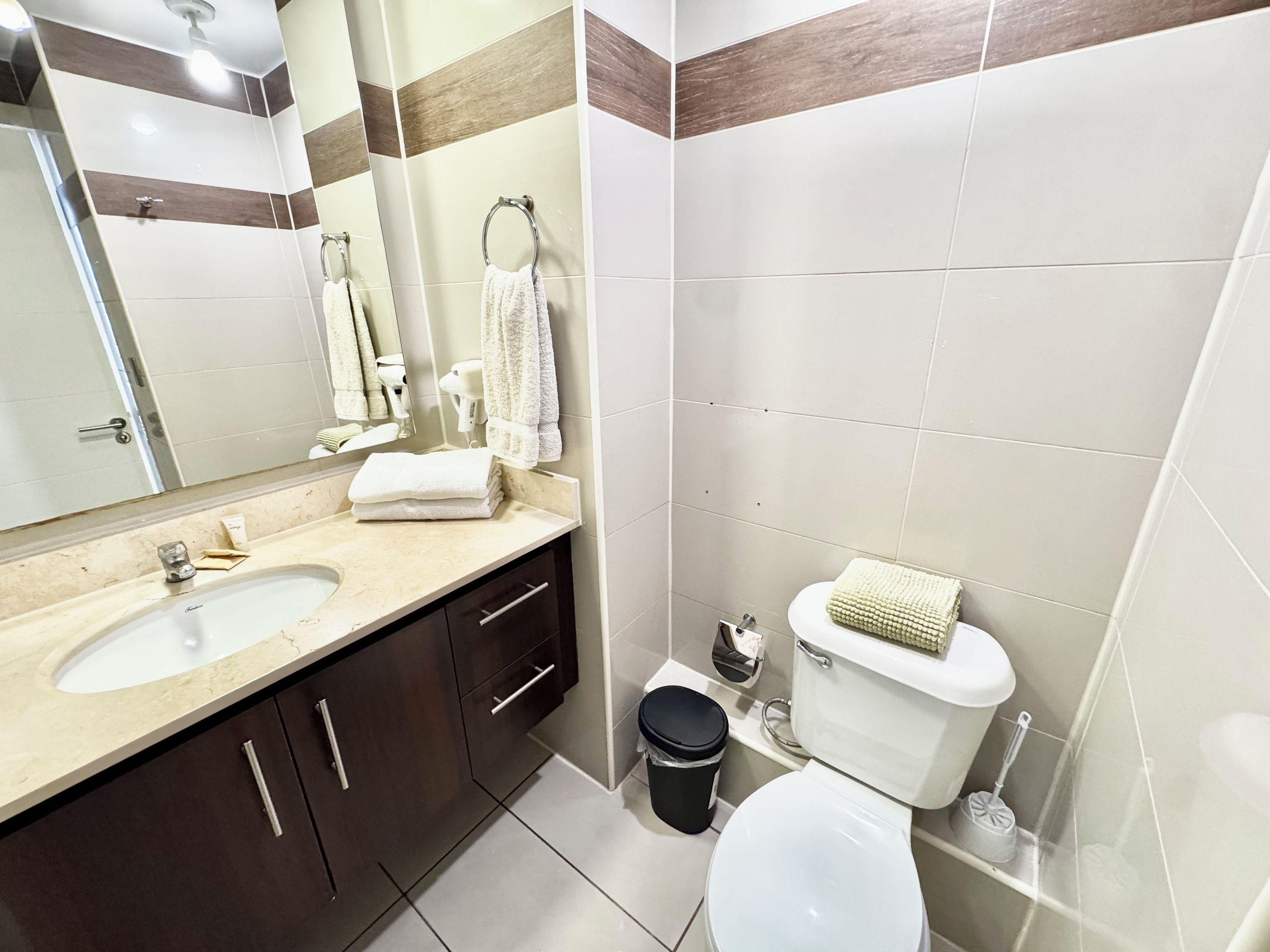 A modern bathroom with two sinks, a toilet, a mirror, and a trash bin.