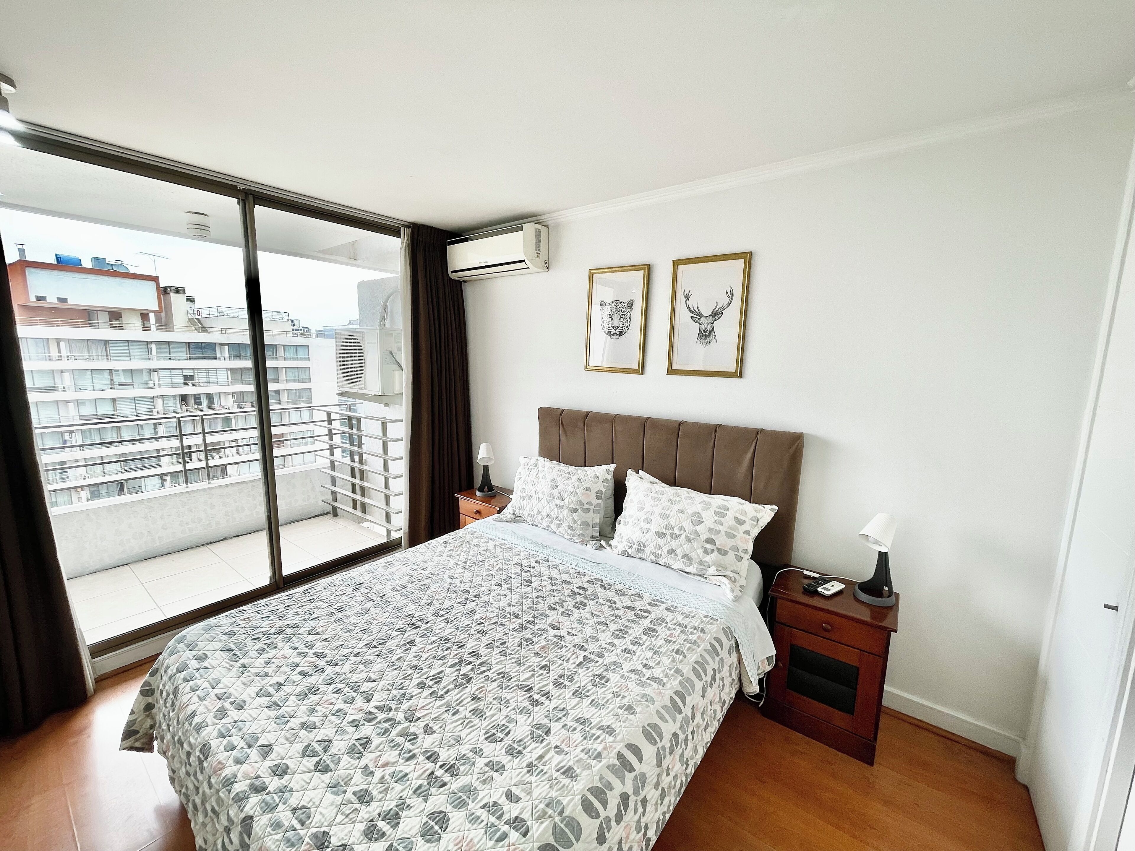 Un dormitorio con una cama grande, una mesita de noche, una ventana con vistas a edificios y dos cuadros en la pared.