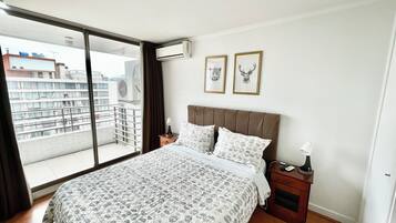 Un dormitorio con una cama grande, una mesita de noche, una ventana con vistas a edificios y dos cuadros en la pared.