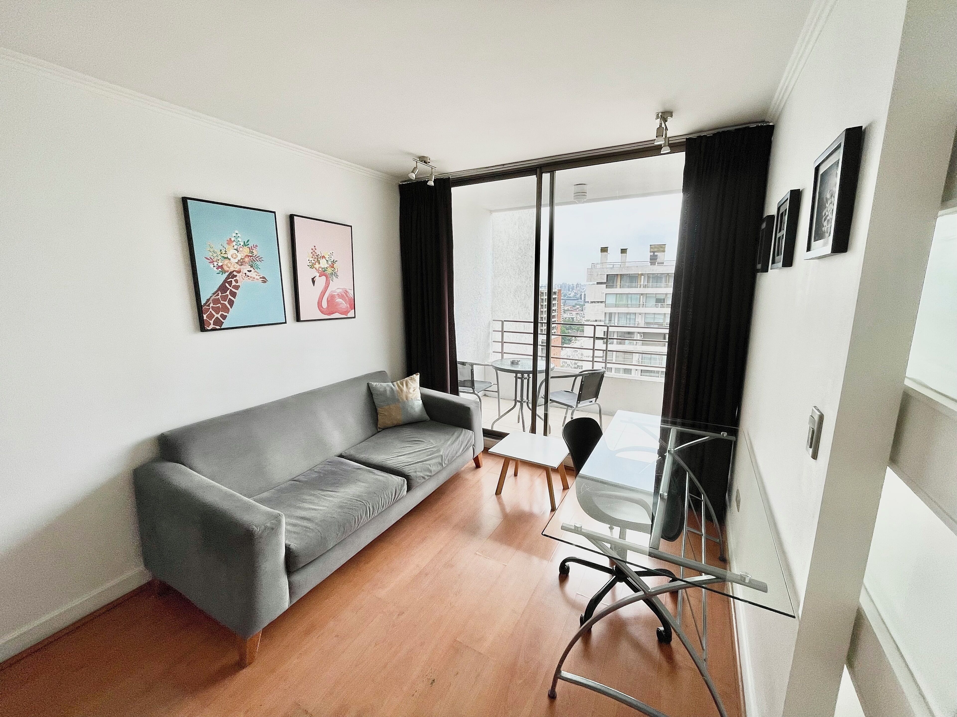 A modern living room with a grey sofa, a glass table, and chairs. There are two framed pictures on the wall.