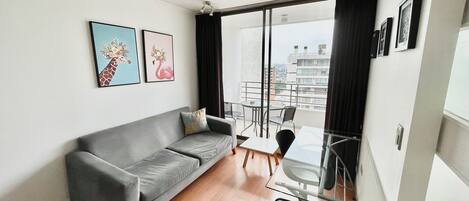 A modern living room with a grey sofa, a glass table, and chairs. There are two framed pictures on the wall.