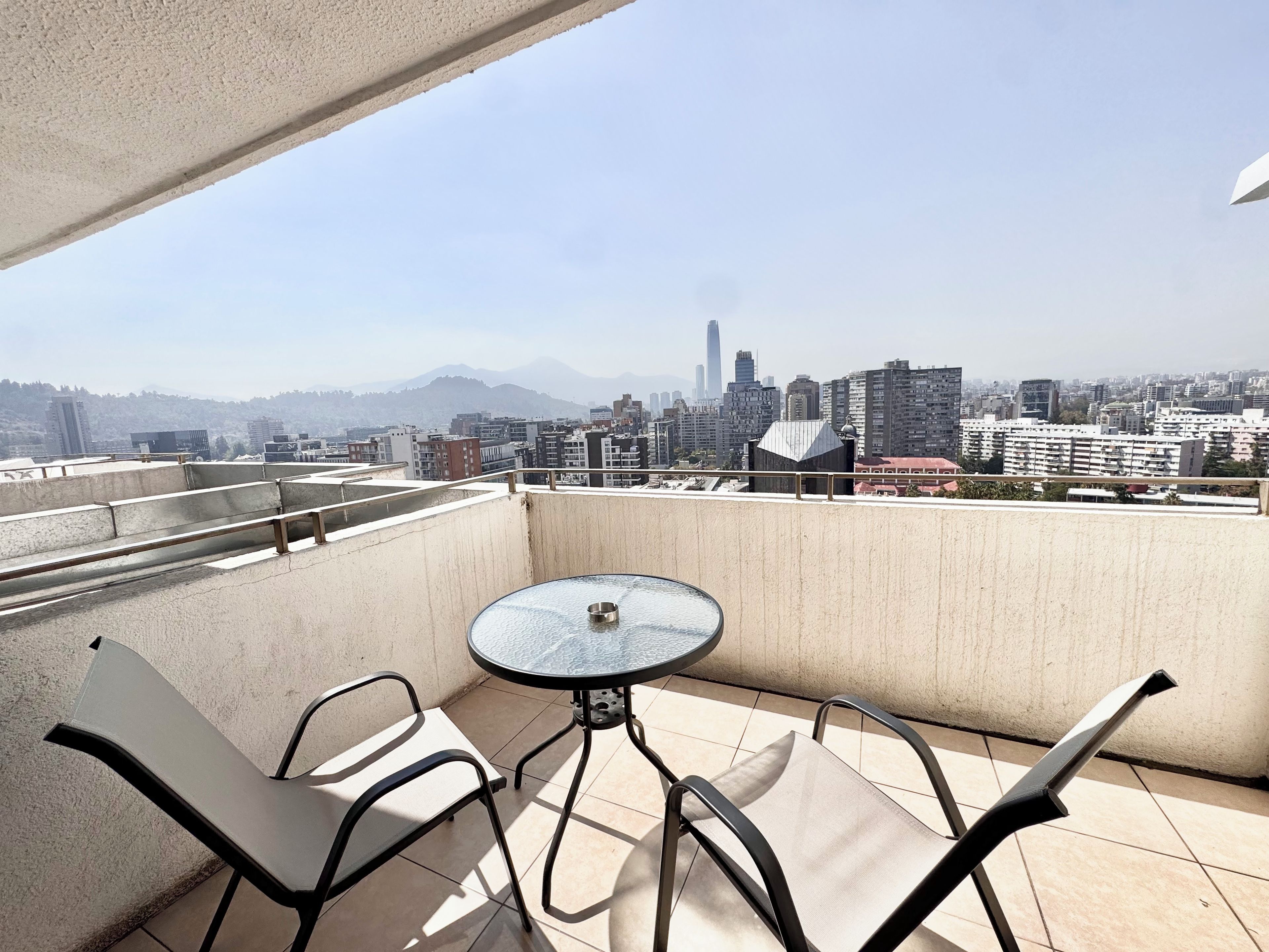 A balcony with a round table and two chairs, overlooking a cityscape with buildings and mountains.