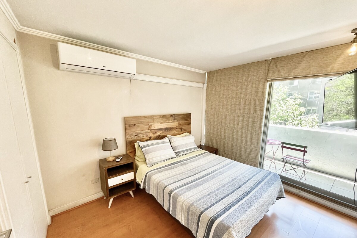 A bedroom with a wooden headboard, a bed with striped bedding, a nightstand with a lamp, and a window with a view of the outside.