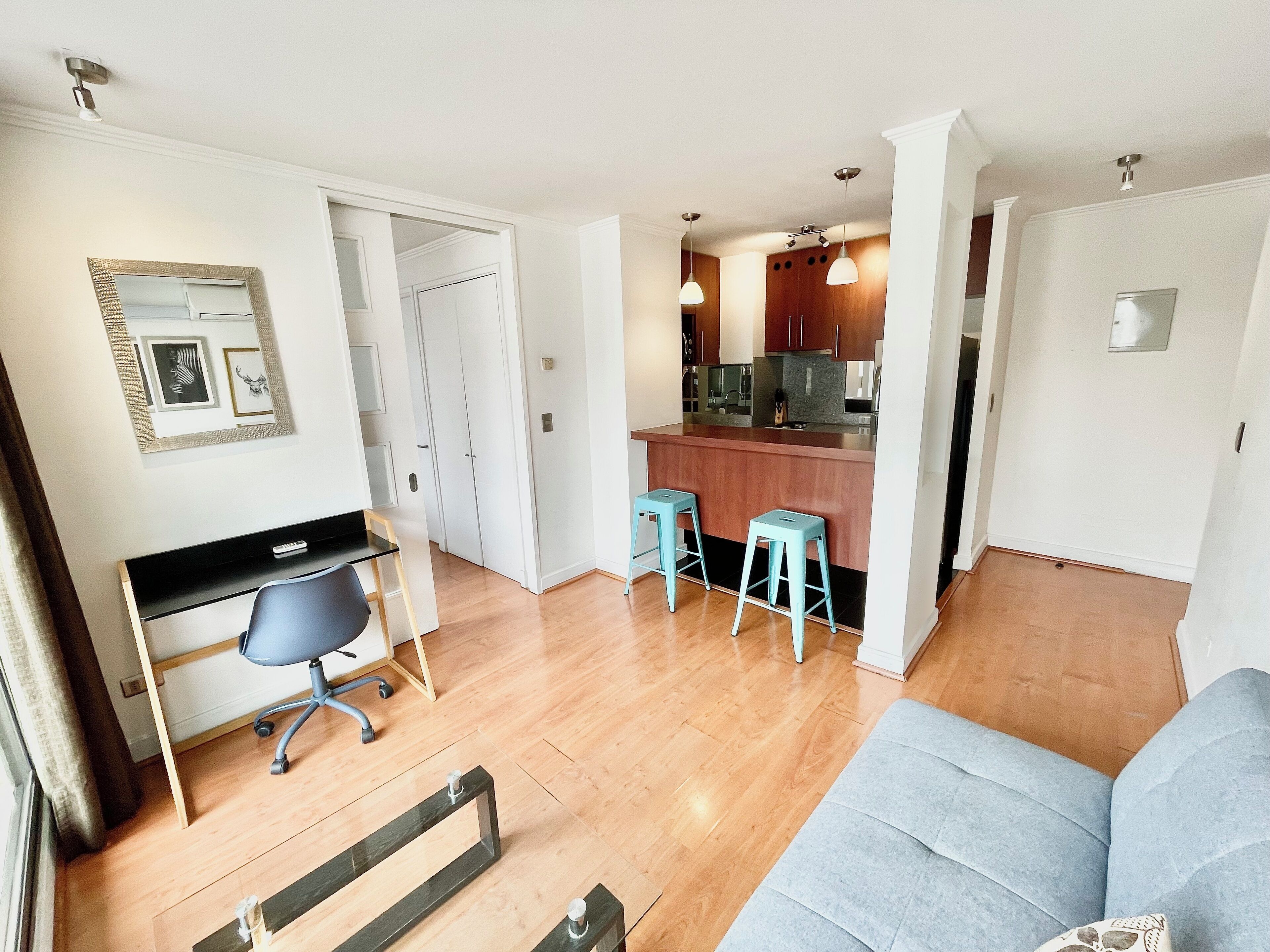 A modern living room with a sofa, a desk with a chair, and a kitchen area with stools.