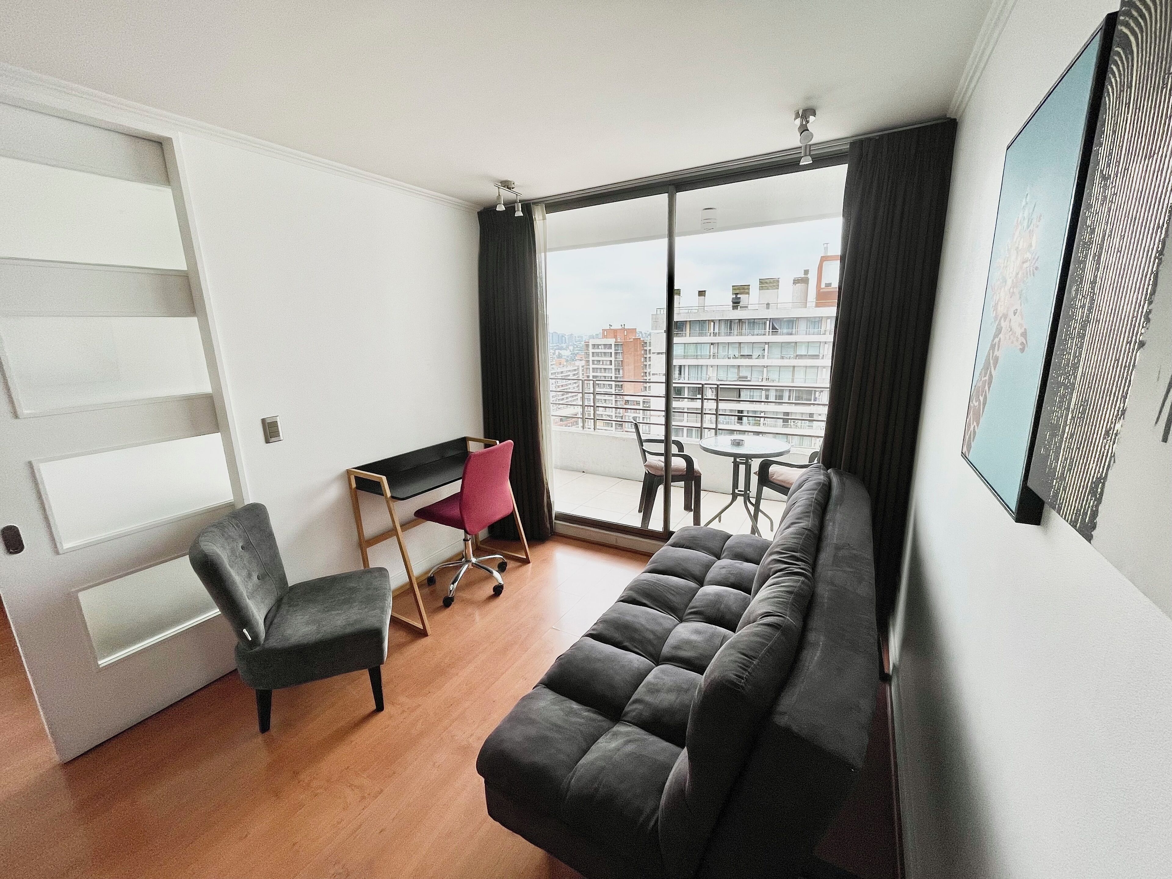 Un salon moderne avec un canapé, un bureau et une chaise. Une grande fenêtre offre une vue sur les immeubles.