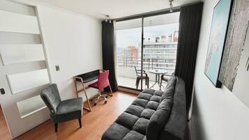 Un salon moderne avec un canapé, un bureau et une chaise. Une grande fenêtre offre une vue sur les immeubles.